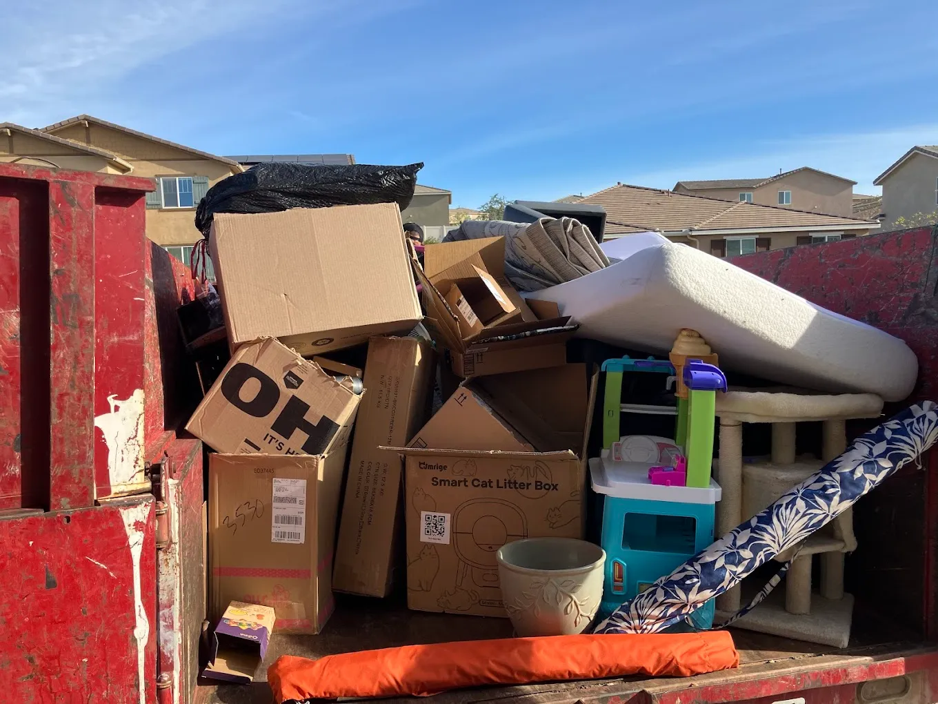Junk Removal Dumpster Filled with Household Items Red junk removal dumpster overflowing with discarded household items including cardboard boxes, a mattress, children's toys, and a cat tree.